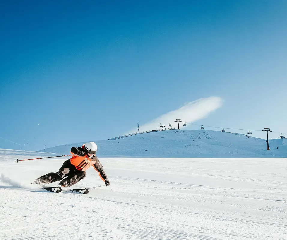 Ein-Skifahrer-in-orange-schwarzer-Skibekleidung-zieht-in-scharfer-Kurvenlage-Spuren-ueber-eine-bestens-praeparierte-sonnenbeschienene-Skipiste-unter-strahlend-blauem-Himmel