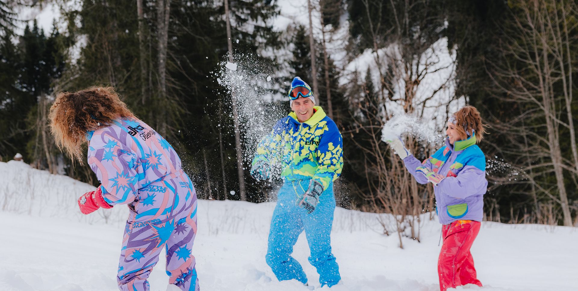 Drei Personen in farbenfroher Skibekleidung werfen sich im verschneiten Wald gegenseitig mit Schnee ab.
