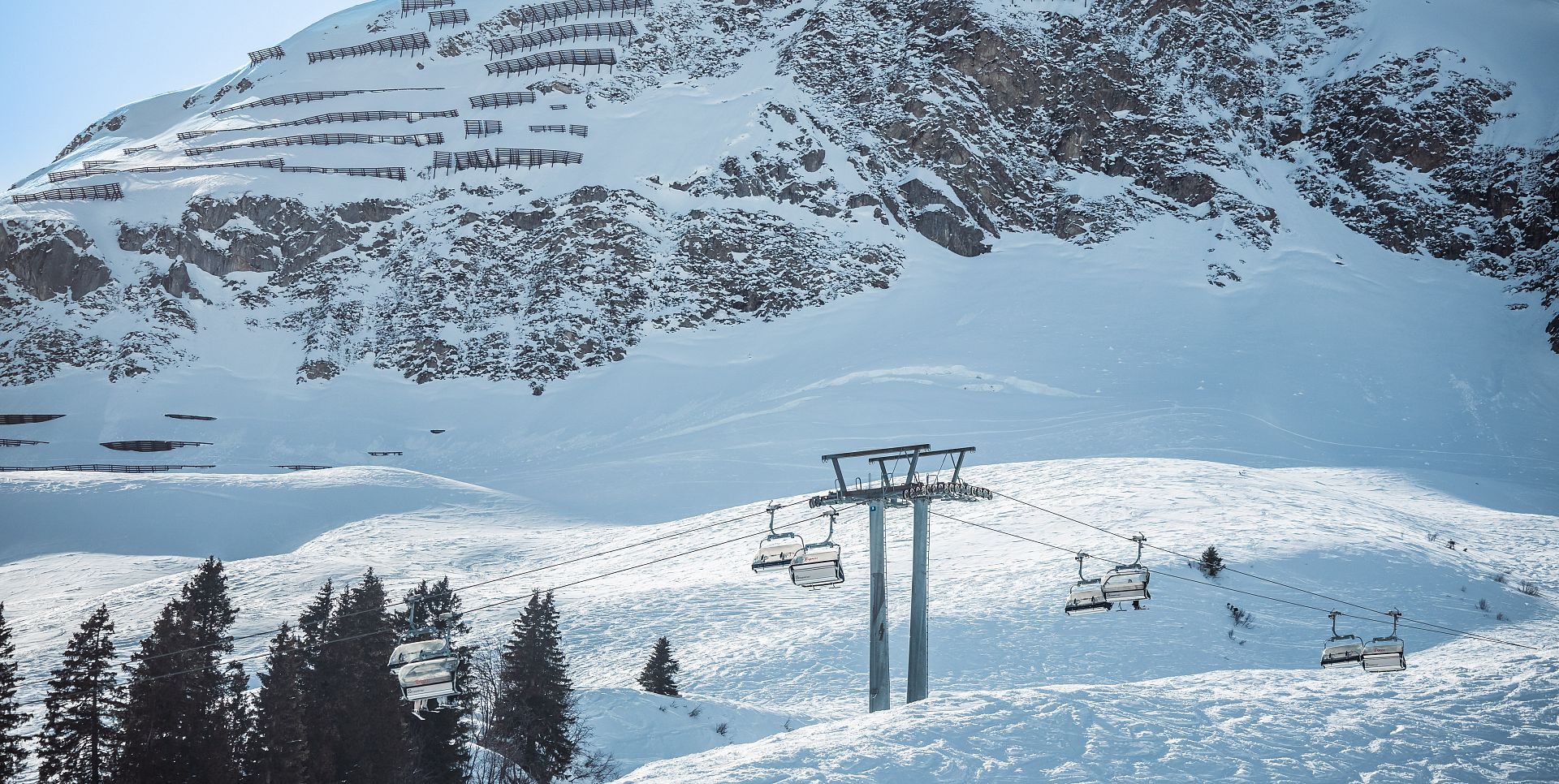 Ein Sessellift befördert Skifahrer über eine verschneite Hanglandschaft vor einem steilen, felsigen Bergmassiv mit Lawinenschutzverbauungen bei sonnigem Wetter.