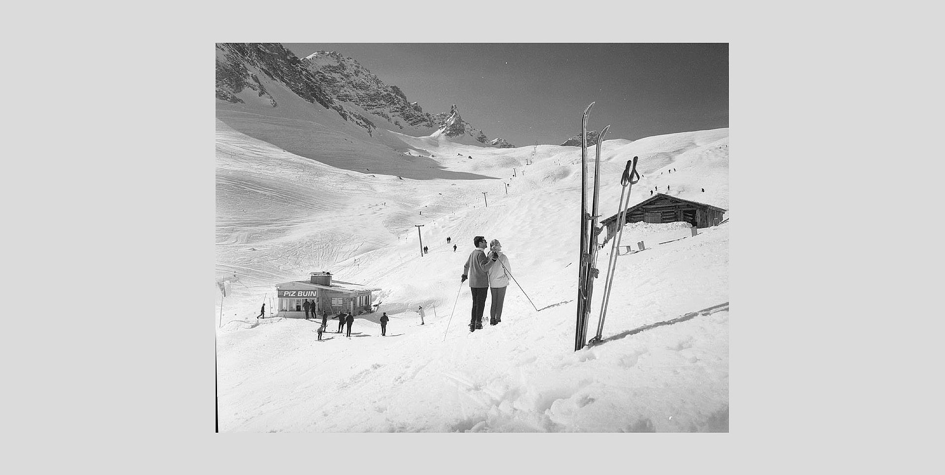 Historic photo of two skiers gazing at the mountains, with the old Wartherhorn Express beside them.