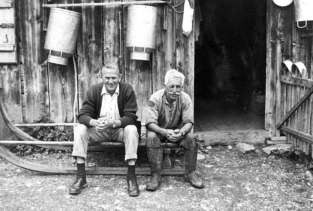 Historic image of two men sitting on a sledge in front of a farmhouse.