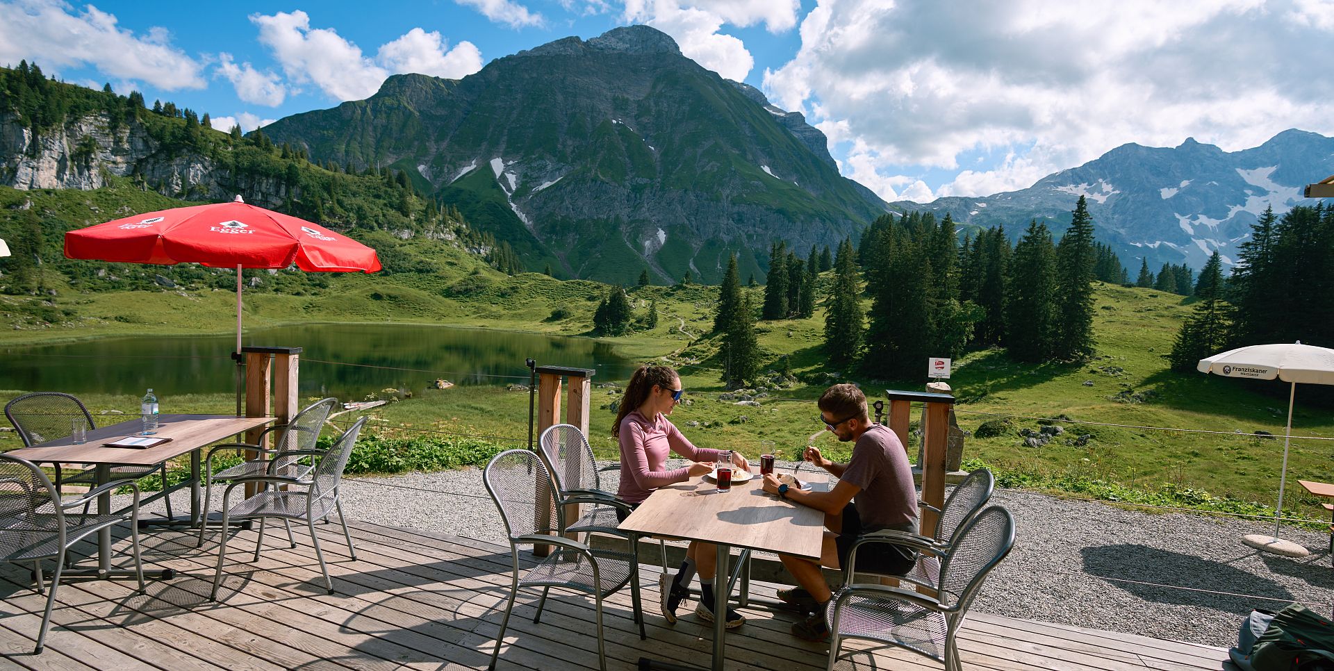 Two people sit on the terrace of the Berghotel Körbersee, enjoying their meal, with the picturesque Körbersee stretching out behind them.