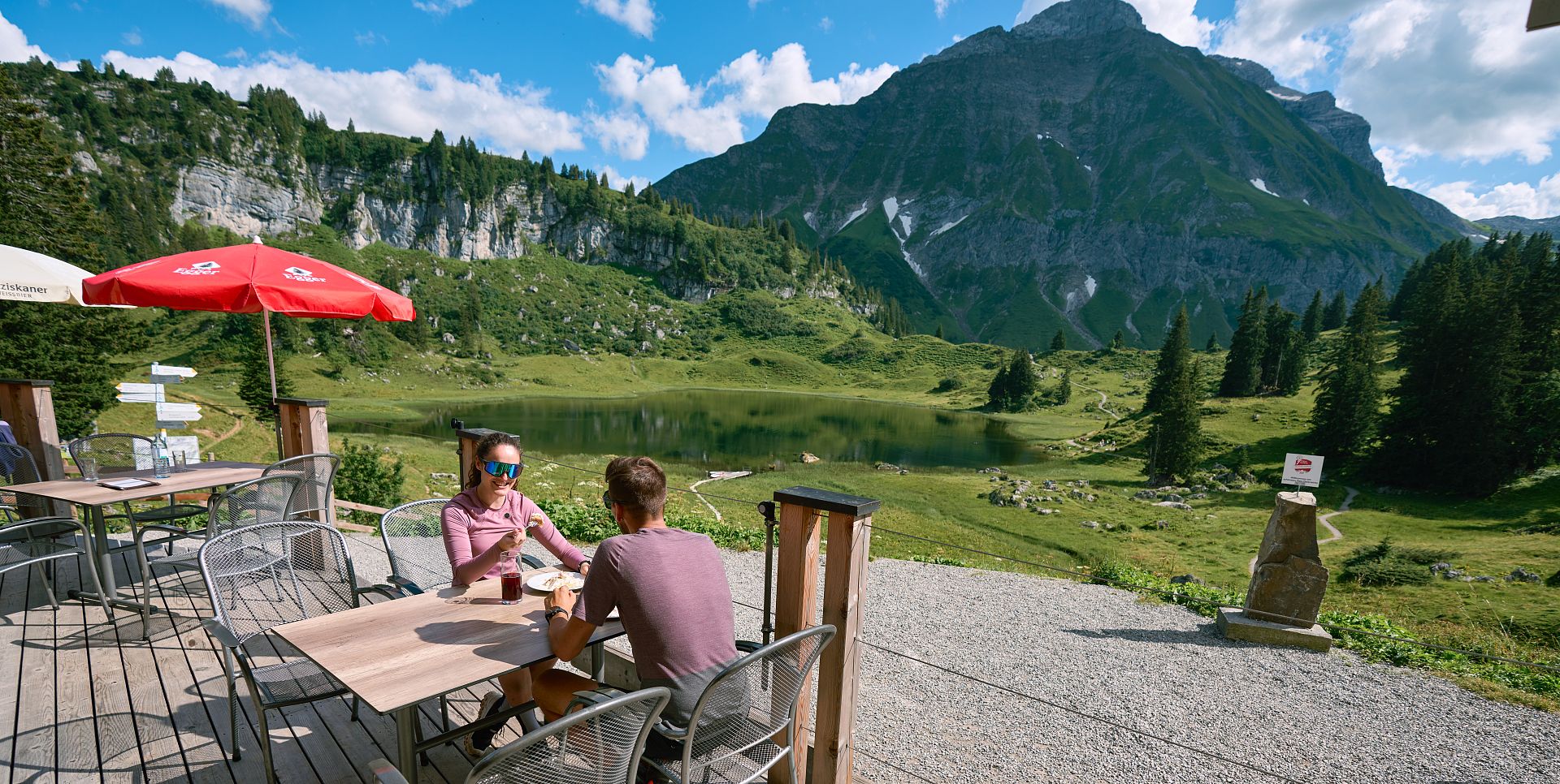 Two people sit on the terrace of the Berghotel Körbersee, enjoying their meal, with the picturesque Körbersee stretching out behind them.