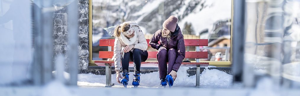 Two women sit on a red bench, putting on ice skates and chatting.