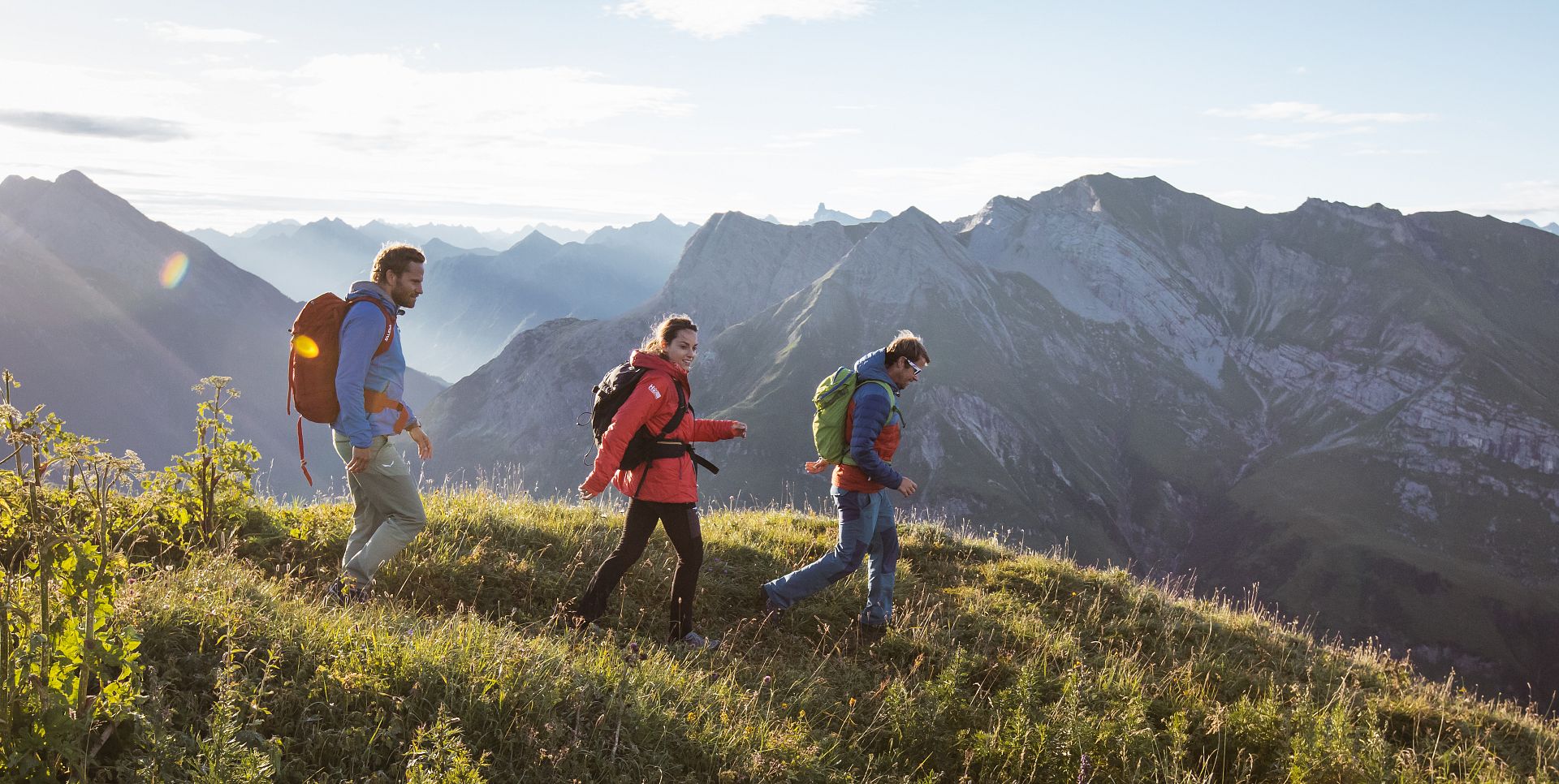 Three people hike across a meadow, with a mountain range stretching out in the background.