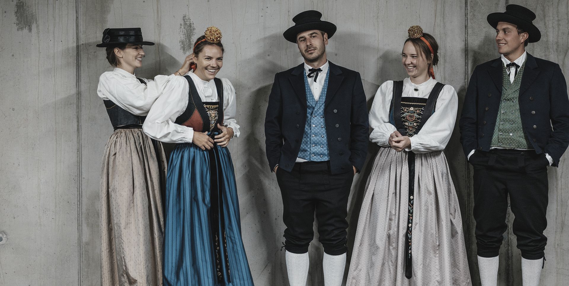 Men and women in traditional Walser costume are standing in front of a concrete wall.