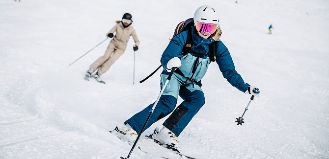 Zwei Skifahrerinnen fahren hintereinander einen Hang hinunter.