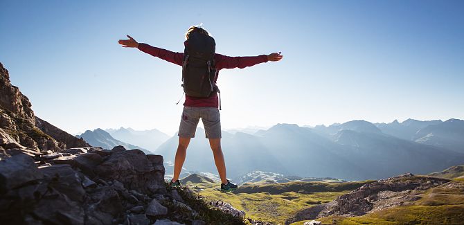 A person stands with arms outstretched, gazing at the surrounding mountain range.