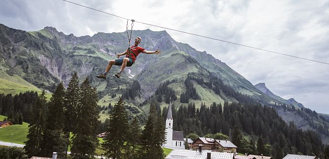Ein Mann schwebt an einer Zipline über das idyllische Bergdorf Schröcken, die markante Kirche im Hintergrund.