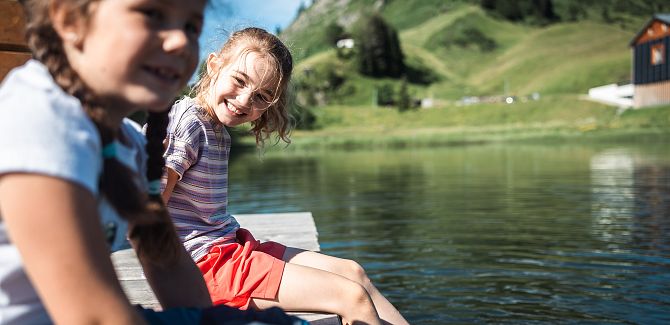 Zwei Kinder sitzen am Ufer und lassen ihre Füße im Seebachsee baumeln.