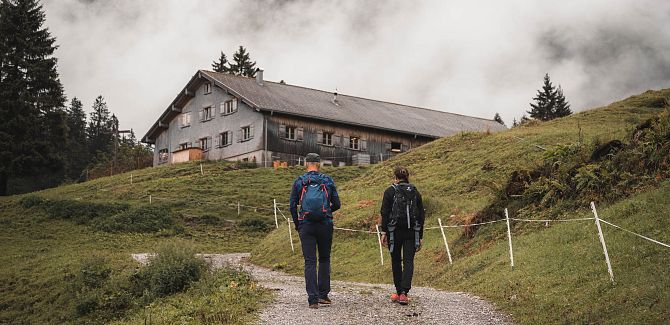 Two people hiking through mist towards an alpine hut in the mountains.