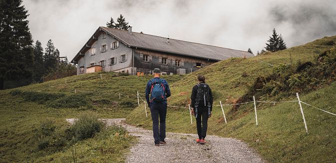 Zwei Personen wandern bei Nebel zu einer Alphütte in den Bergen.