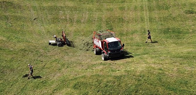 Johannes and Hubert Strolz haymaking. Shot from above – visible is a fully mown slope with a tractor loaded with hay and a mower. Two people can also be seen, small in the frame, holding hay rakes.