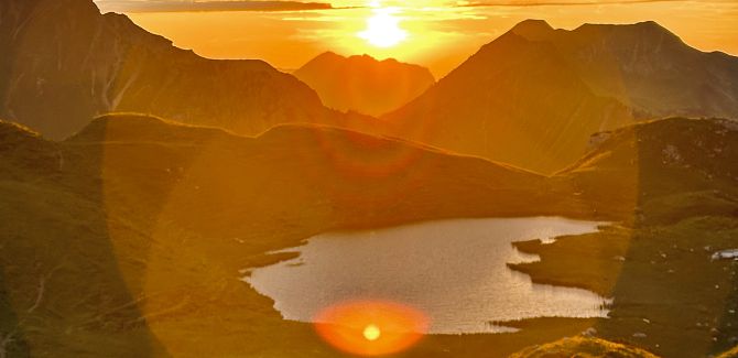 Der Hochalpsee liegt ruhig, während hinter ihm der Himmel in einem leuchtenden Orange des Sonnenuntergangs erstrahlt.