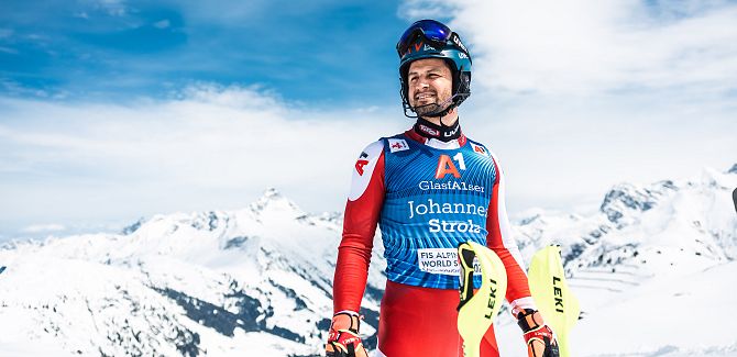 Johannes Strolz stands in his racing suit, with the snow-covered mountains behind him.