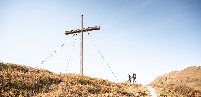 The Weltfriedenskreuz on the Simmel, with autumnal landscape in the foreground, blue sky in the background, and two hikers approaching the cross.