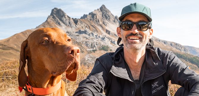 A portrait of Andi Tomann sitting in the meadow, with a mountain range visible behind him. His dog sits beside him.