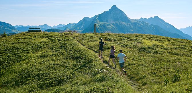 Three trail runners run up to a bench, with the Biberkopf visible in the background.