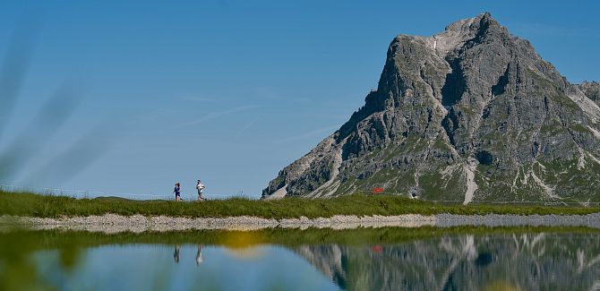 Two trail runners run along Panorama Lake, with the Widderstein rising in the background.