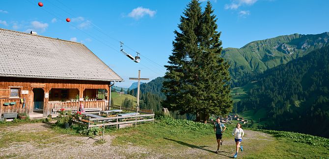 Two trail runners pass by Alpe Felle, with the material cable car visible in the background.