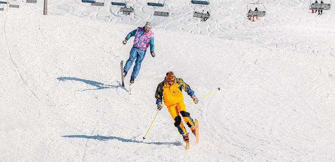 Zwei Skifahrende in farbenfroher Kleidung fahren gemeinsam eine verschneite Piste hinunter.
