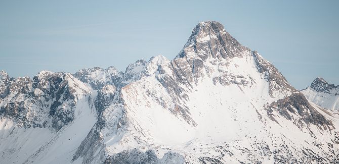 Ein imposanter, schneebedeckter Felsgipfel ragt vor einem hellen, leicht bewölkten Himmel über eine schroffe alpenländische Gebirgskette empor.