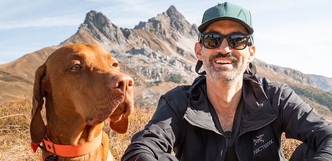 A portrait of Andi Tomann sitting in the meadow, with a mountain range visible behind him. His dog sits beside him.