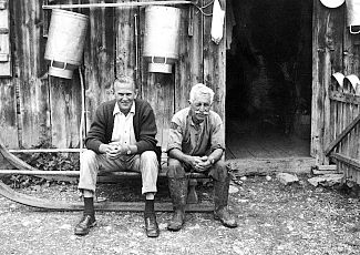 Historic photo of two men sitting on a sled in front of a farmhouse.
