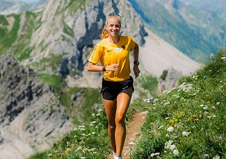Eine Trailrunnerin in einem gelben Shirt läuft in Richtung Wartherhorn, während im Hintergrund die Berglandschaft zu sehen ist.