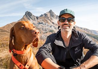 A portrait of Andi Tomann sitting in the meadow, with a mountain range visible behind him. His dog sits beside him.