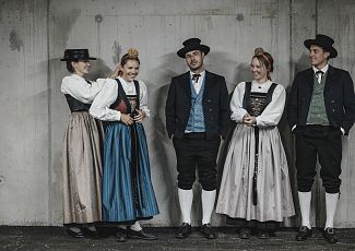 Ladies and gentlemen in Walser traditional costume pose in front of a concrete wall.