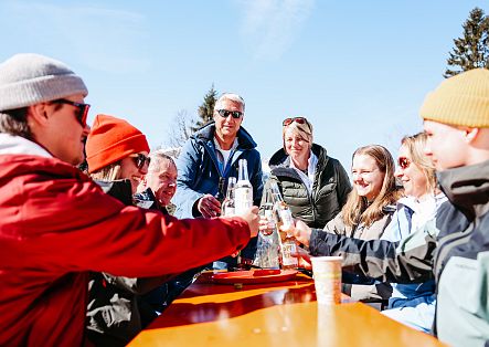 Eine Gruppe von Menschen steht im Freien um einen Tisch und stößt mit Getränken an.