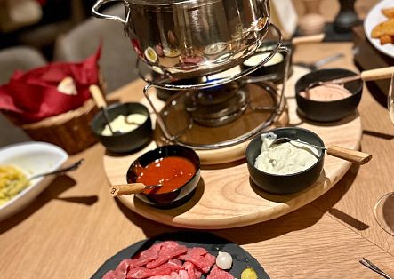 Ein gedeckter Holztisch mit einem Fondue-Set im Zentrum, umgeben von kleinen Schälchen mit Saucen, Dips und rohem Fleisch.