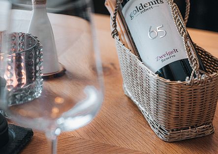 Eine Flasche Rotwein in einem kleinen Weidenkorb sowie ein Weinglas auf einem elegant gedeckten Tisch.