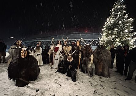Eine Gruppe von Krampussen steht vor einem festlich geschmückten Christbaum, gemeinsam mit dem heiligen Nikolaus und Engeln.
