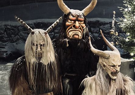 Drei furchteinflößende Krampusse stehen bedrohlich vor einem festlich geschmückten Christbaum. Sie tragen imposante, detailreiche Masken mit langen Hörnern, wildem Fell und grimmigen Gesichtszügen.