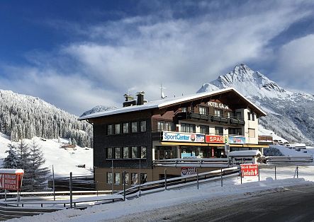 Verschneites Sportgeschäft mit Skiverleih an einer Bergstraße, dahinter eine schneebedeckte Berglandschaft.