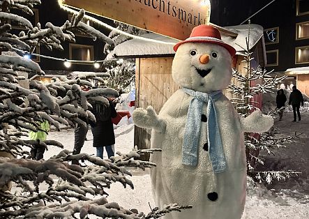 Ein großer Schneemann mit rotem Hut und blauem Schal steht zwischen verschneiten Tannen auf einem Weihnachtsmarkt.