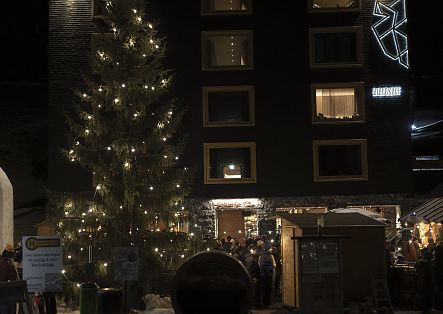 Ein beleuchteter Weihnachtsbaum und Marktstände stehen vor einem dunklen Gebäude auf dem verschneiten Dorfplatz.