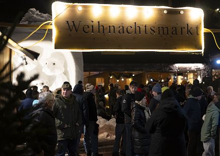 Zahlreiche Menschen besuchen abends einen verschneiten Weihnachtsmarkt mit beleuchteten Verkaufsständen.