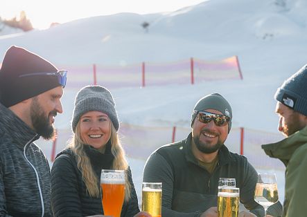 Vier Personen sitzen im Freien vor einer verschneiten Berglandschaft und trinken gemeinsam.