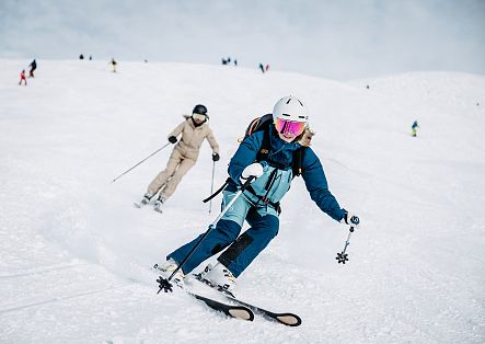 Eine Person in blauer Skibekleidung fährt mit Skiern eine verschneite Piste hinunter, dahinter folgt eine weitere Person.
