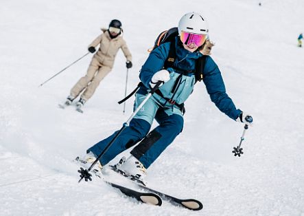Eine Skifahrerin fährt in einer Kurve über eine verschneite Piste, während dahinter eine weitere Person folgt.