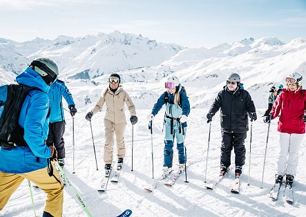 Mehrere Skifahrende stehen gemeinsam auf einem verschneiten Berg und blicken auf die umliegenden Gipfel.