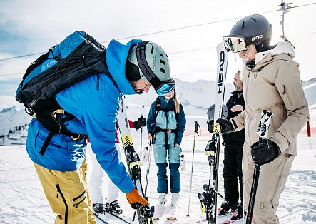 Eine Gruppe von Skifahrenden steht im Schnee und richtet ihre Skiausrüstung, während ein Skilehrer sie anleitet.