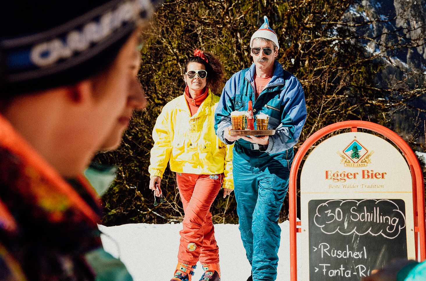 Zwei Personen in farbenfroher Skibekleidung tragen ein Tablett mit Getränken durch den Schnee.