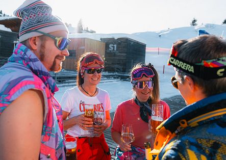 Vier Personen stehen in Skibekleidung im Schnee zusammen und unterhalten sich mit Getränken in der Hand.