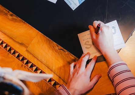 Eine Person schreibt mit einem Stift in ein kleines Notizbuch auf einem Holztisch.