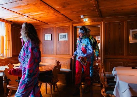 Zwei Personen in Skibekleidung betreten eine holzvertäfelte Gaststube, während eine weitere Person am Tisch steht.