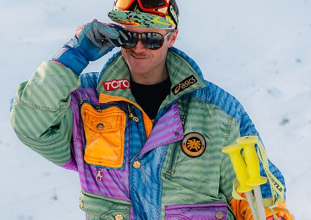 Ein Skifahrer mit farbenfroher Bekleidung hält seine Skibrille hoch und blickt in die Ferne.